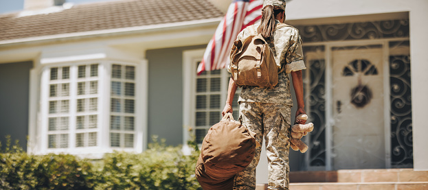 Back view of a military mom holding a teddybear while standing outside her house with her luggage. Courageous female soldier coming back home after serving her country in the army.