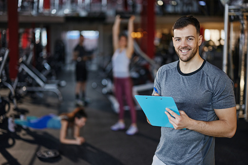 Happy fitness instructor writing sports training plans in a gym and looking at camera. There are people in the background.