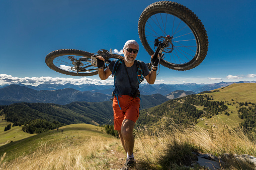 A middle aged male mountainbiker is carrying his mountainbike on shoulders on a sunny summerday in Weissensee region of Carinthia, Austria. The region is very famous in wintertime for ice skating and in summertime for hiking and mountainbiking.\nCanon EOS 5D Mark IV, 1/1600, f/3,2 , 16 mm.