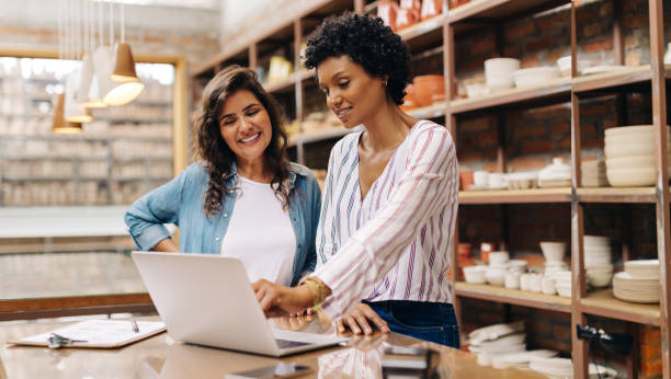 Successful female ceramists using a laptop together in their store Successful female ceramists using a laptop while working together. Two female entrepreneurs managing online orders in their store. Happy young businesswomen running a creative small business together. art and craft product stock pictures, royalty-free photos & images