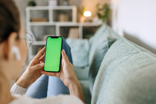 Young woman using phone with green screen while relaxing on the sofa