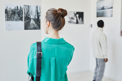 Back view of young woman wearing green while looking at art in gallery or museum, copy space