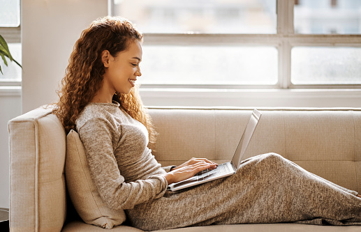 Woman with laptop on sofa typing on keyboard for online search on internet, networking or surfing ecommerce website in her living room or home apartment. Young person on couch check email app online