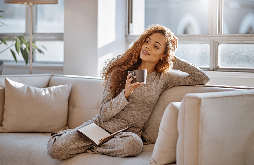 Woman drink coffee on sofa to relax in living room comfortable, smile and happiness at home in morning. Calm Brazil girl happy relaxing, tea and reading notebook on lounge couch at house or apartment