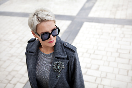 Portrait of fashionable mature woman with short blonde hair wearing stylish gray wool coat with lapels and shoulder straps, accessories brooch and sunglasses