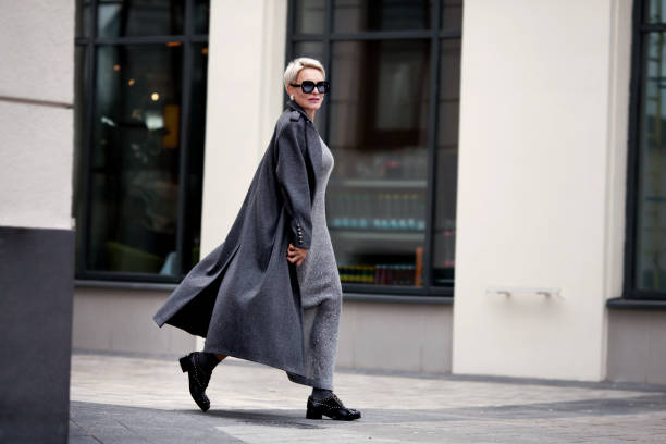 elegante donna matura cammina per strada in città, indossa abiti eleganti, cappotto di lana grigia, abito lungo, scarpe e occhiali neri. outfit di tendenza - single lane road foto e immagini stock