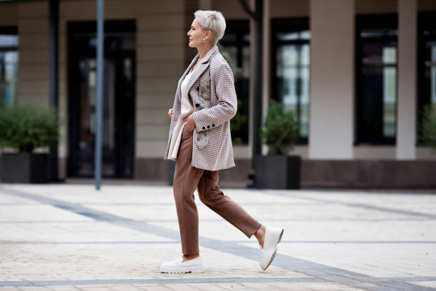 Side view full length woman walks outside and wears trendy business clothes, oversize jacket, tie, pants and white loafer shoes. Confident female model with short blonde hair stock photo