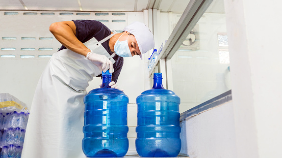 man worker in workwear and with a protective mask on his face working in a drink water factory checking blue water gallons before shipment.