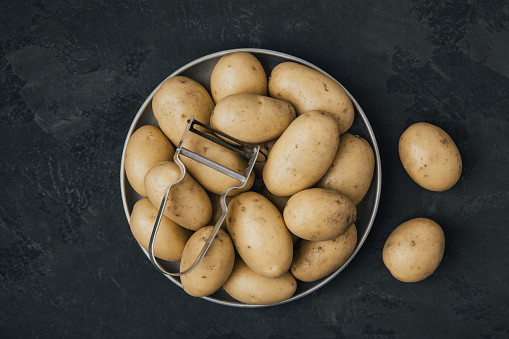 Potato. Fresh raw organic potatoes in bowl with peeler on dark stone background. Top view with copy space.