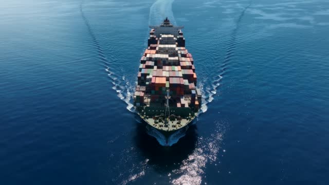 Aerial top view of a large container cargo vessel in motion passing by over blue, calm ocea