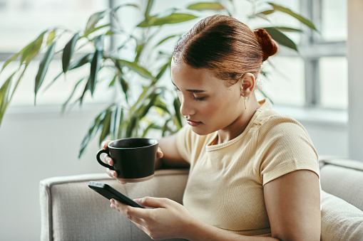 Woman, phone and coffee of a person from Atlanta with technology on a living room lounge sofa. Social media, mobile and web scroll of a young female on a home couch using internet at a house