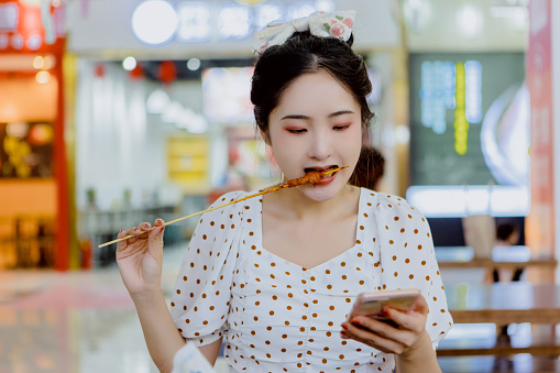 A woman eating barbecue kebabs playing mobile phone in a mall enjoying nightlife