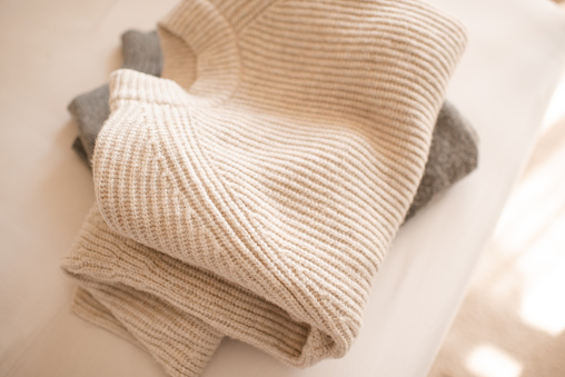 Stack of knitted wool textile sweaters clothes on white blanket in bed at home room close up. Winter cozy season.