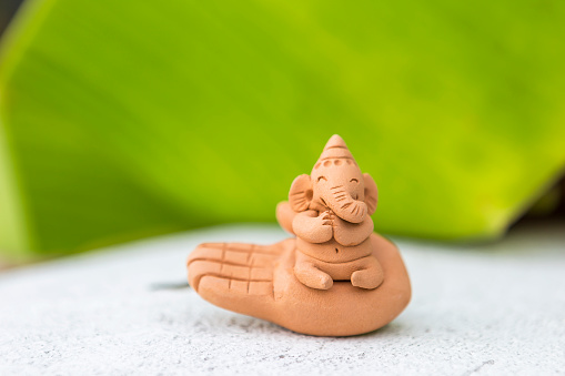 Cute little Ganesh clay sculpture with space on blurred green banana leaf background, Indian god, culture and religion