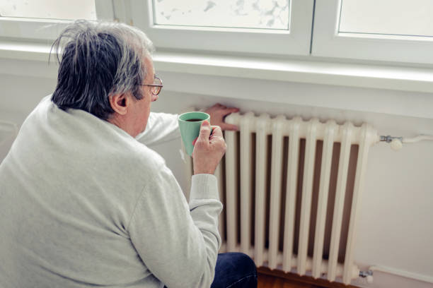 Dissatisfied senior men having problem with central heating, sitting on sofa at home, freezing, drinking hot tea trying to warm up. Energy crisis concept.