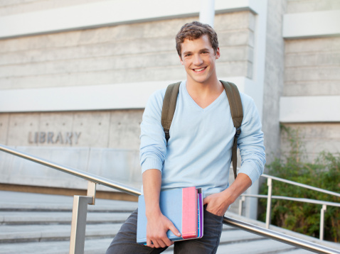 A young handsome male Caucasian student is walking on the campus carrying books in his arms with headphones around his neck with his friends talking in the background.