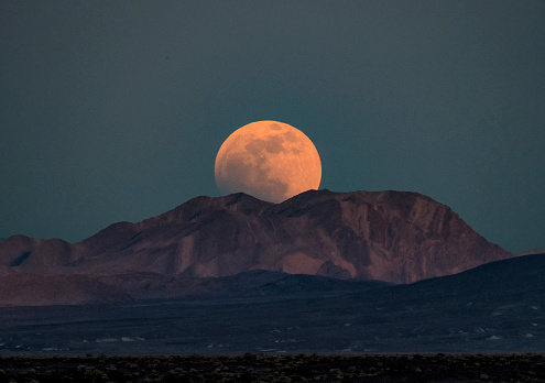A Strawberry Super Moon is rising behind a mountain range at Death Valley Junction, California. the telephoto image captures the exceptionally large moon as it clears the mountains.