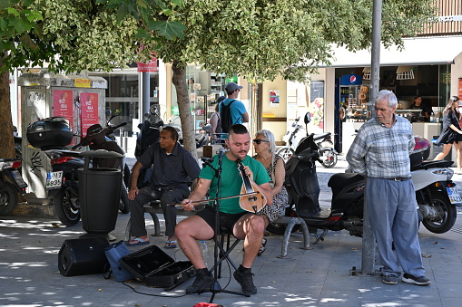 Heraklion, Greece - 22 09 2022: Street musician singing and playing Cretan lyra, traditional Greek music instrument in centrum of town surrounded by people. He wears green t-shirt and black shorts.