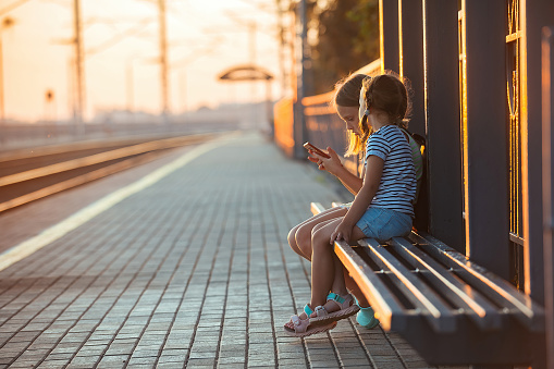 two little girls tourist sisters are sitting on the bench of the railway station and watching the phone while waiting for the electric train in the evening in the rays of the setting sun