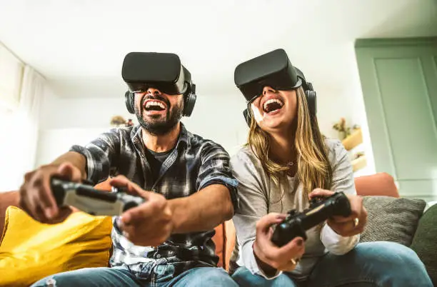 Happy friends sitting on sofa playing video games with virtual reality glasses - Cheerful couple having fun with new trendy technology videogames - Hobby and tech concept