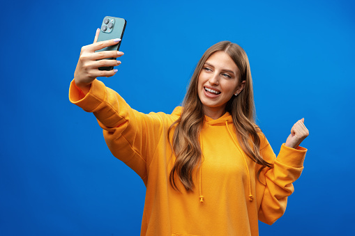 Photo of attractive young woman takes selfie photo on smartphone on blue background in studio