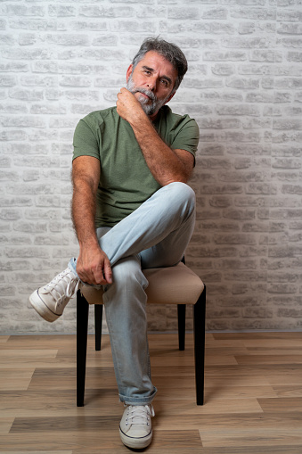 Casually handsome. Studio shot of handsome mature man sitting on the chair and looking at camera with smile, vertical. Portrait of a trendy guy sitting on chair against white brick wall, vertical