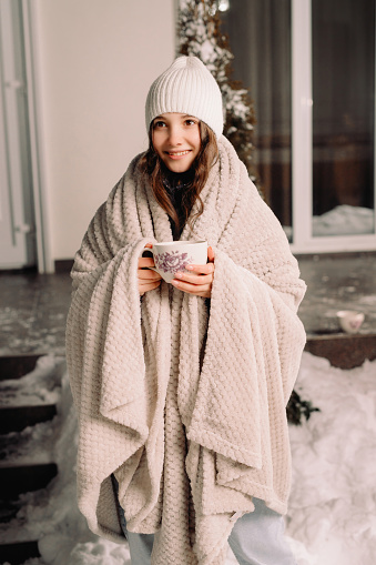 Full-length photo of a beautiful happy little girl wearing white hat and blanket holding a cup of hot tea outside on winter evening. . High quality photo