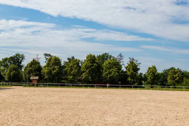 Photo of Fence for horses where races are held. There is sand on the ground, trees and blue sky with white clouds on the background.