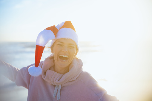 Portrait of happy modern middle aged woman in cosy sweater with striped christmas hat having fun time at the beach in the evening.