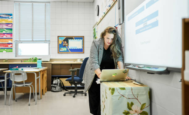School teacher using a laptop and a projector for a classroom lesson School teacher preparing for a lesson using a screen projector and a laptop while working at a desk in her classroom teacher projector stock pictures, royalty-free photos & images