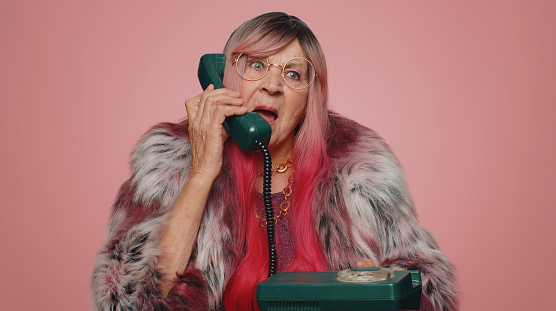 Crazy tourist hacker elderly old woman talking on wired vintage telephone of 80s, fooling, making silly faces. Senior stylish grandmother granny hotline agent isolated alone on pink studio background