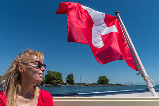 Mature blonde woman with sunglasses smiling and Swiss flag fluttering on the stern of a boat on Lake Geneva.