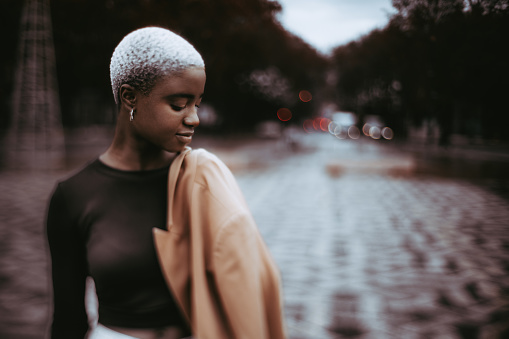 A true tilt-shift low-key portrait with a shallow depth of field and a selective focus on a part of the face of a youthful cute black female standing in the middle of a city boulevard next to the road