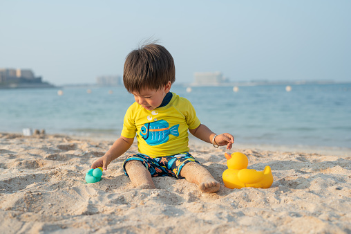 Adorable one year , 14 months old mixed race baby playing with a toy duck on the beach siting on the sand and enjoying the seaside breeze in an unrecognizable tourist resort at sunset. Mixed race infant boy playing with toys in the sand on the beach