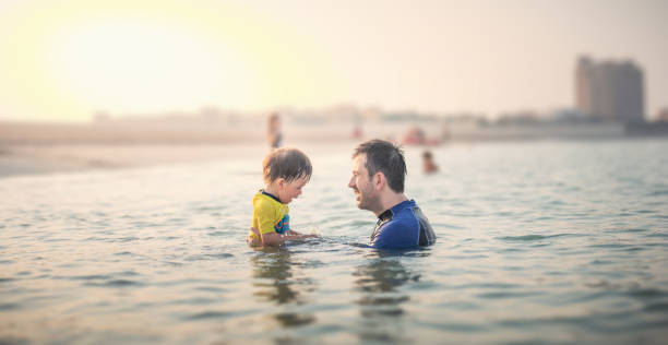 Father and infant son having fun at the beach in the shallow seaside water at sunset stock photo