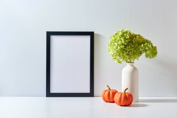 istock Mockup with a black frame and green flowers in a vase, pumpkins on a light background. Empty poster frame mockup for presentation design, text, lettering 1430794463