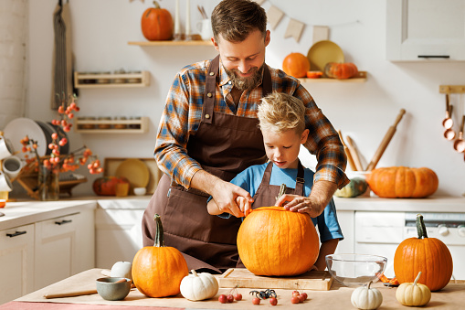 Cute happy little boy helping his father to carve Halloween pumpkin while standing in kitchen at home and preparing for autumn holiday, family son and dad making Jack-o-Lantern together