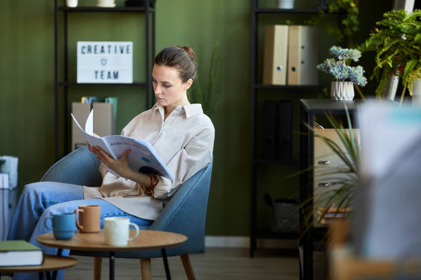 Young economist in casualwear sitting in armchair and looking through manual Young manager or economist in casualwear sitting in comfortable armchair and looking through manual in openspace office handbook stock pictures, royalty-free photos & images