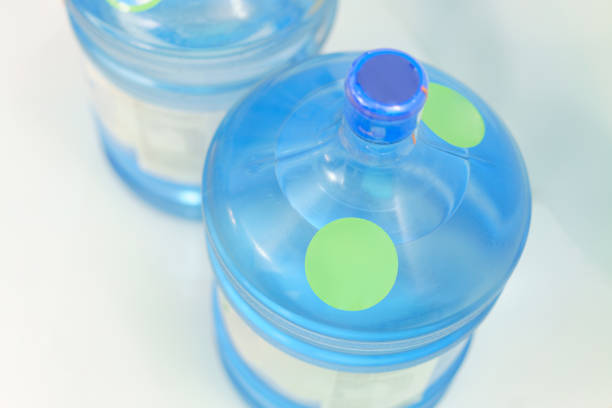 The top of two plastic gallon water bottle containers with green round labels stand on a blue background. Drink. Beverage. Large. Pure. Full. Plastic. Big. White. Delivery. Liquid. Barrel. Container la parte superior de dos recipientes de botellas de agua de galón de plástico con etiquetas redondas verdes se encuentra sobre un fondo azul. beber. bebida. grande. puro. lleno. plástico. grande. blanco. entrega. líquido. barril. contenedor