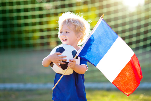 Kids play football on outdoor field. France team fans with national flag. Children score a goal at soccer game. Child in French jersey and cleats kicking ball. Fan celebrating victory at pitch.