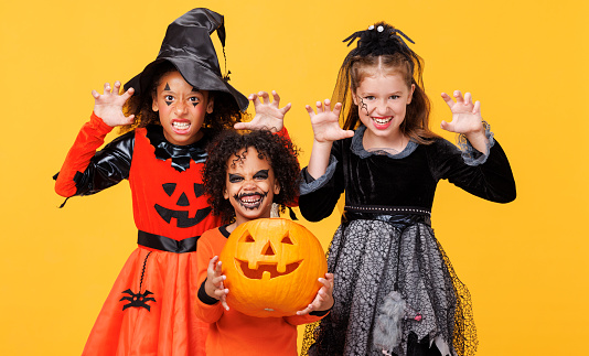 Happy Halloween! Cheerful kids in carnival costumes and makeup make a terrible gesture on bright colored yellow background