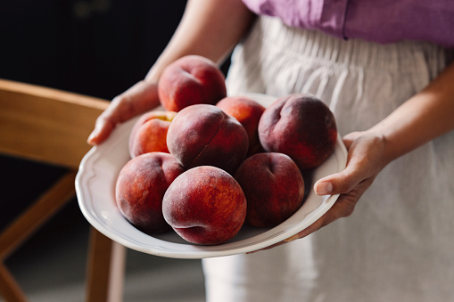 A cropped photo of an anonymous Caucasian woman holding a plate of fresh juicy peaches.
