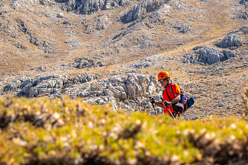 60 years old man. He is trekking. Mountainous extreme region. Orange t-shirt, backpack, walking pole, helmet