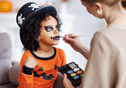 Festive makeup for Halloween. Woman doing pumpkin make-up  for cheerful ethnic curly boy in costume while preparing holiday at home.