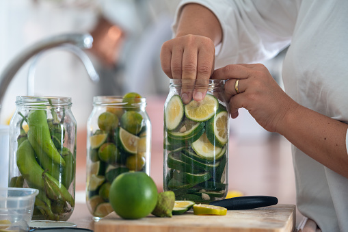 Photo of woman hand preparing fruit and vegetables for seasoning. Limes, lemons, oranges and green peppers are seen in jars. Shot from a low angle viewpoint with a full frame mirrorless camera in kitchen.