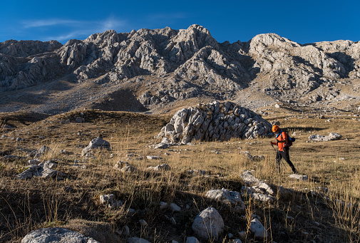 60 years old man. He is trekking. Mountainous extreme region. Orange t-shirt, backpack, walking pole, helmet.Steep mountain in blue clear sky background.