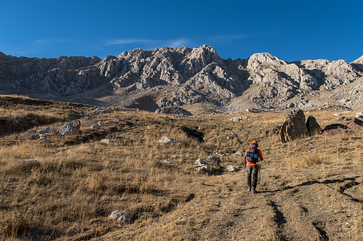 60 years old man. He is trekking. Mountainous extreme region. Orange t-shirt, backpack, walking pole, helmet.Steep mountain in blue clear sky background.