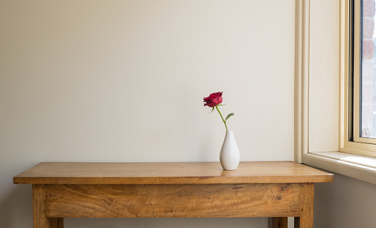 Single red rose in white vase on oak side table against beige wall next to window with sunlight