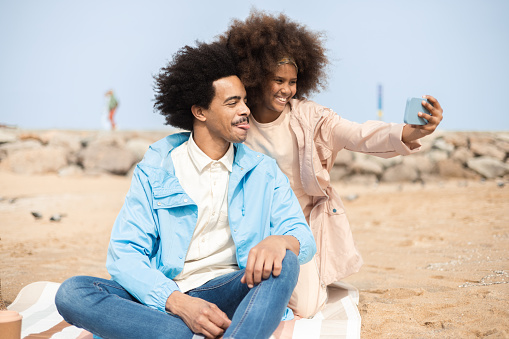 Portrait of a happy African American man and his teenage daughter enjoying a picnic on the beach and taking a selfie