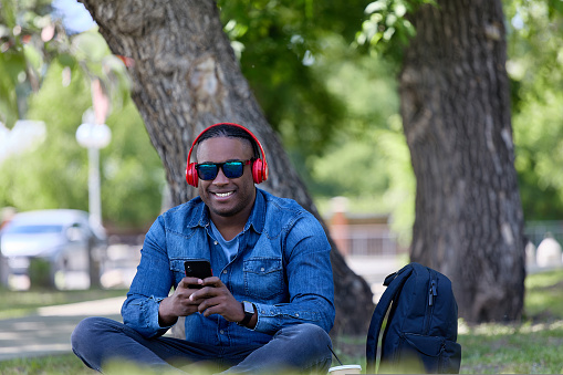 Photo freelancer man enjoying nature while working outdoors using a mobile phone. Stylish African student studies a subject online while spending his free time in the park